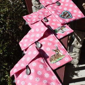 Puede incluir: Cuatro bolsas de tela rosa con patrones florales blancos y detalles bordados. Cada bolsa tiene un cierre de botón plateado y presenta un diseño bordado diferente, que incluye un corazón con la palabra "Schatz", un ciervo, una escena de montaña y un corazón con la palabra "Herz".