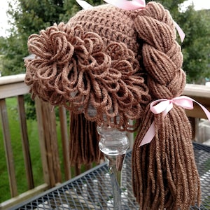 May include: A brown crocheted hat featuring yarn hair styled into a braided ponytail and bangs. The hat is adorned with light pink satin ribbons tied at the base of the braid and at the top of the crown, creating a decorative hair-like appearance.