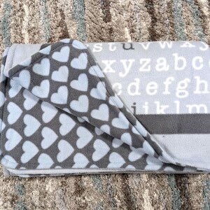May include: A folded gray and blue fleece blanket. One side features a pattern of white hearts on a gray background. The other side is gray with white letters spelling out the alphabet. A dark gray stripe runs across the middle.