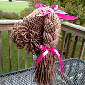 May include: A brown crocheted hat featuring yarn hair styled into two thick braids and a side bun. The hair is accented with bright pink satin ribbons tied at the top of the braids and at the base of the long, loose ends of the braided yarn hair.