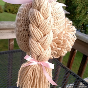 May include: A beige crochet hat featuring a thick, braided yarn ponytail and a textured side section. The hat is adorned with two light pink satin ribbons, one tied at the top of the braid and another securing the base of the yarn ponytail.