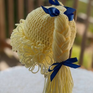 May include: A pale yellow crocheted hat featuring a textured fringe along the front edge and a long, thick yarn braid attached to the side. The braid is secured at the top and bottom with dark blue satin ribbons tied into small bows.
