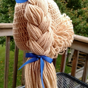 May include: A beige crocheted hat featuring a thick, braided yarn ponytail and a side bun made of looped yarn. The hairstyle is accented with two bright blue satin ribbons, one tied at the top of the braid and one at the base of the ponytail.