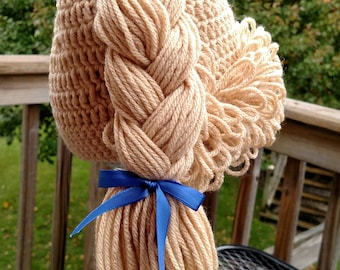 May include: A beige crocheted hat featuring a thick, braided yarn ponytail and a side bun made of looped yarn. The hairstyle is accented with two bright blue satin ribbons, one tied at the top of the braid and one at the base of the ponytail.