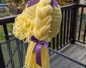 May include: A yellow crocheted hat with long blonde yarn braids and purple satin bows. The hat is displayed on a glass stand.