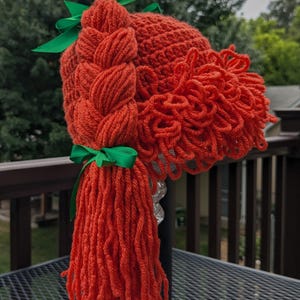 May include: A crocheted hat made of orange yarn, featuring a thick braided side ponytail and a fringe of curly yarn bangs. The hat is accented with two bright green satin ribbons tied at the top and at the base of the braided ponytail.