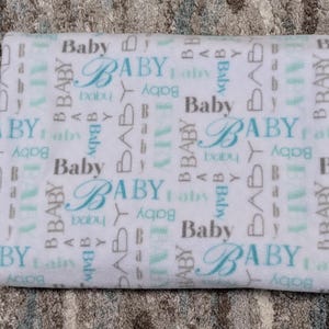 May include: A folded white baby blanket with the word "Baby" repeated in various fonts and colors. The word "Baby" is in a light blue script font, and other instances are in gray and light blue block letters.
