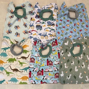 May include: Six baby bibs with various patterns, including dinosaurs, farm animals, construction vehicles, and cartoon dogs. Each bib has a green or gray neck opening and is made of soft fabric.