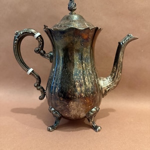 May include: Antique silver-plated coffee pot with ornate handle and spout. The pot has a decorative lid with a finial and rests on three small feet. The exterior shows signs of age and use, adding to its vintage charm. A classic piece for serving coffee or tea.