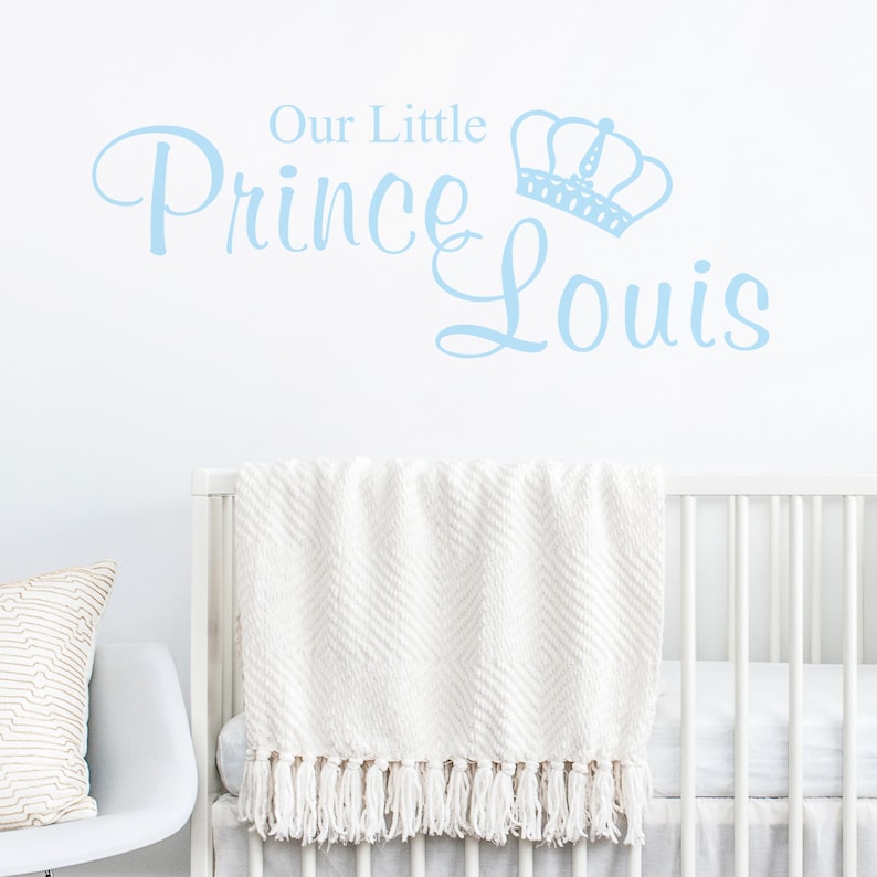 May include: A light blue wall decal reading "Our Little Prince Louis" with a crown illustration. The decal is above a white cot with a white fringed blanket. A white chair with a pillow is on the left.