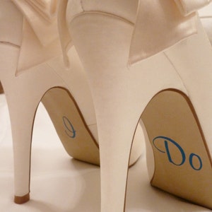 May include: A pair of white satin high-heeled shoes with bows on the toes. The heels are decorated with blue lettering, one shoe says "I Do" and the other says "I Do".