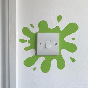 Può includere: Un interruttore bianco con un interruttore rettangolare è centrato su un muro bianco. L'interruttore è circondato da un design a spruzzi di vernice verde brillante. Il muro è accanto a un telaio della porta bianco.
