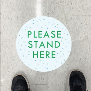 May include: A round floor sticker with the words "PLEASE STAND HERE" in green on a light blue background, decorated with confetti. The sticker is on a grey and white tiled floor, with a person's feet and shoes visible.