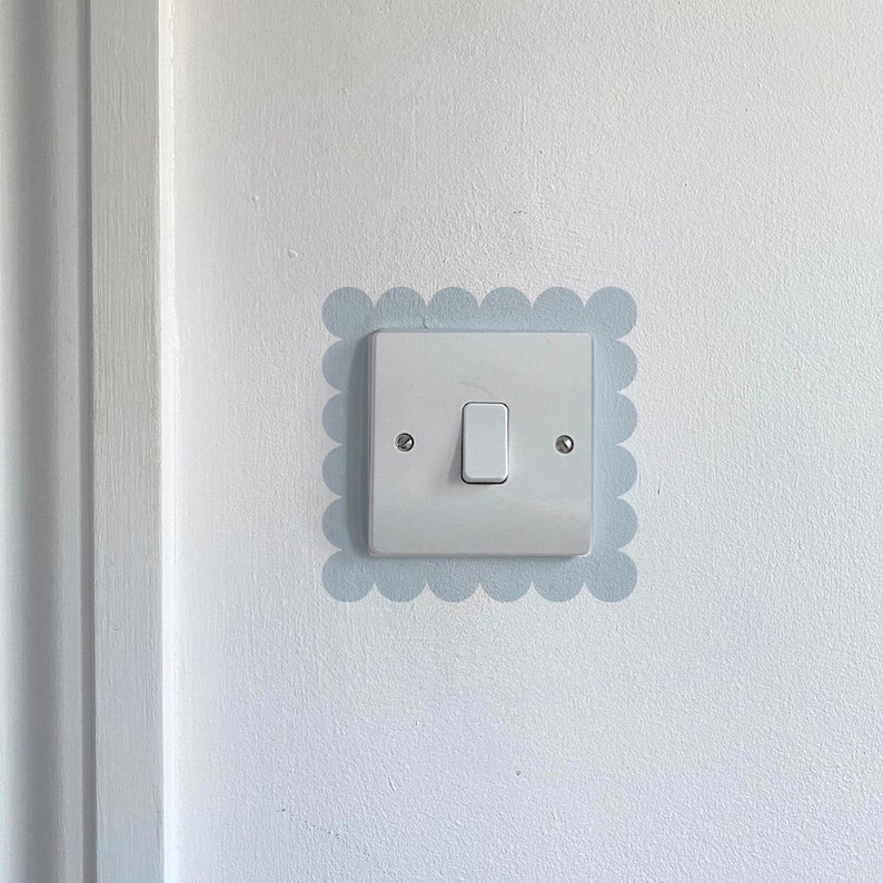 K&ouml;nnte beinhalten: Ein wei&szlig;er Lichtschalter mit einer rechteckigen Platte und einem einzigen Schalter. Der Schalter befindet sich innerhalb eines hellblauen, wellenf&ouml;rmigen Rahmens an einer wei&szlig;en Wand. Zwei kleine Schrauben sind auf der Platte sichtbar.
