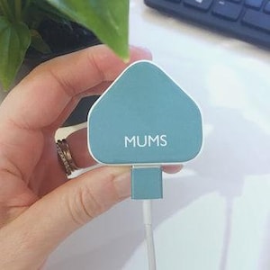 May include: A teal and white charger with the word "MUMS" printed on the front. The charger is held by a hand with a ring on the finger. A white cord extends from the bottom of the charger.
