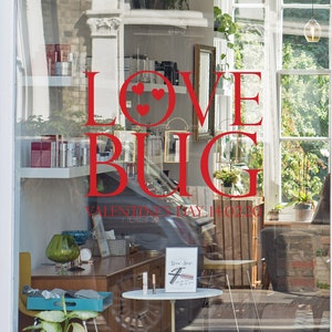 May include: A shop window display featuring the words "LOVE BUG" in red, with heart symbols. The window showcases beauty products and a sign reading "Valentine's Day 14.02.20". The interior is visible through the glass.