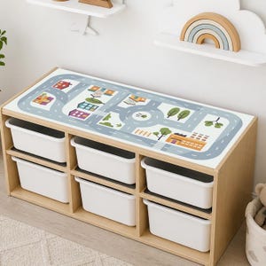 May include: A light-colored wooden toy storage unit featuring six white rectangular plastic bins arranged in two rows. The top surface of the unit displays a printed graphic of a town map with roads, buildings, and trees in muted colors.