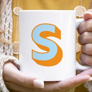 Pode incluir: Caneca de cerâmica branca com uma grande letra "S" estilizada. A letra é azul clara com uma sombra laranja, criando um efeito 3D. A caneca é segurada por uma pessoa.