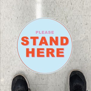 以下が含まれることがあります： 「PLEASE STAND HERE」の文字がオレンジとピンクで書かれた、水色の円形のフロアステッカー。赤い縁取りがある。画像の下部には黒いスニーカーが見える。床はグレーです。