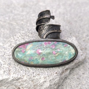 Könnte beinhalten: Ein handgefertigter Silberring mit einem ovalen, türkis gefärbten Stein, der mit rosa Punkten eingelegt ist. Das Band des Rings hat ein strukturiertes, gewickeltes Design. Der Ring liegt auf einer grauen, strukturierten Oberfläche.