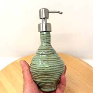 Pode incluir: Um dispensador de sabonete de cerâmica feito à mão com um design texturizado e canelado em tons de verde e castanho. Possui uma bomba e um bico de aço inoxidável. O dispensador é segurado em uma mão.