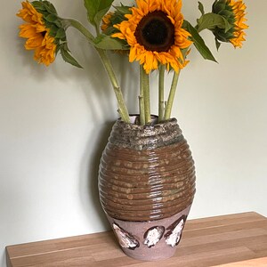 May include: A brown ceramic vase with a textured surface and white accents holds a bouquet of yellow sunflowers.