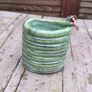 May include: A green ceramic planter shaped like a coiled snake with a red tongue sticking out. The planter is sitting on a wooden surface.