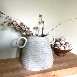 May include: A white ceramic vase with two handles and horizontal blue lines, containing dried flowers. A small brown bowl filled with seashells sits nearby on a wooden surface, creating a coastal theme.