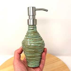 Op de afbeelding: Een keramische zeepdispenser met een getextureerd, geribbeld ontwerp in groen- en bruintinten. De dispenser heeft een roestvrijstalen pomp en tuit. De fles wordt in een hand gehouden, wat de grootte en vorm laat zien.