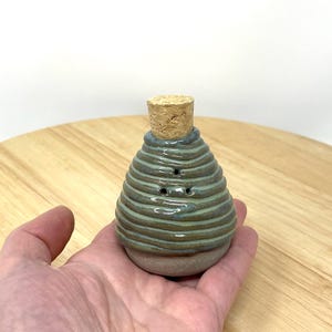 Op de afbeelding: Een kleine, handgemaakte keramische zout- of peperstrooier met een kurken dop. De strooier is geglazuurd in een lichtblauwe en bruine kleur met een gestreept patroon.