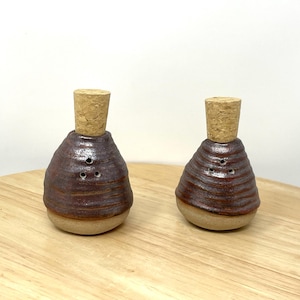 May include: Two brown ceramic salt and pepper shakers with cork tops. The shakers have a textured, rippled design and three holes on the top for dispensing salt and pepper.