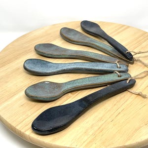 Pode incluir: Um conjunto de seis colheres de cerâmica em tons de azul, verde e preto, cada uma com um pequeno orifício para pendurar. As colheres estão dispostas numa tábua redonda de madeira, mostrando sua aparência artesanal única.