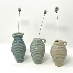 Puede incluir: Tres jarrones de cerámica con un diseño texturizado y acanalado. Los jarrones son en tonos azules y beige. Cada jarrón tiene una pequeña abertura en la parte superior y una asa en un lado.