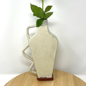 May include: White ceramic vase with a unique, twisted handle. The vase has a speckled finish and a red base.