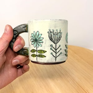 以下が含まれることがあります： 濃い色の取っ手とクリーム色のボディのセラミックマグ。マグカップは、手描きのターコイズと黒の花柄で装飾されています。ハンドルは、質感のある濃い緑色の仕上げです。マグカップは手で持たれています。