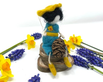 May include: A felt doll wearing a yellow hat and blue dress sits on a wooden base with a pine cone in its hand. The doll is surrounded by yellow daffodils and blue flowers.