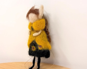 May include: A handmade felt doll with a tan face, brown hair, and a yellow coat over a black dress. The doll has small black legs and a white ear. The doll is standing on a wooden surface.