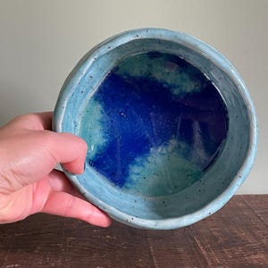May include: A light blue ceramic bowl with a speckled glaze. The inside of the bowl is glazed in a blue and green color.