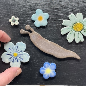 Könnte beinhalten: Eine Sammlung von Keramikblumen- und Schnecken-Dekorationen in den Farben Blau, Grün und Gelb. Die Blumen sind in verschiedenen Formen und Größen, und die Schnecke ist hellbraun.