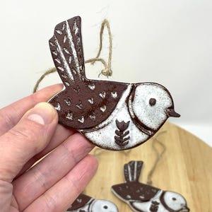 Puede incluir: Un adorno de pájaro de cerámica con esmalte marrón y blanco. El pájaro tiene un pequeño diseño de flor en el pecho y una cuerda para colgar.