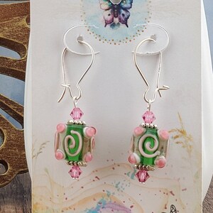 Può includere: Un paio di orecchini d'argento con perline di vetro rosa e verde. Le perline hanno un design a spirale e sono decorate con cristalli trasparenti. Gli orecchini sono presentati su una carta bianca con il testo "Tranquil Journey" e un indirizzo di sito web.