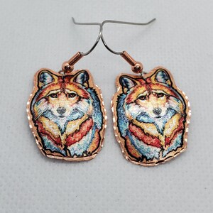 May include: A pair of copper earrings featuring a colorful wolf head design. The wolf's fur is depicted in shades of red, orange, yellow, blue, and white. The earrings have a silver hook closure.