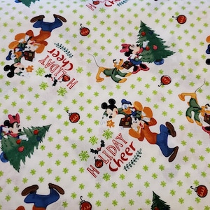 Op de afbeelding: Witte stof met een herhaald patroon van Disney-figuren, kerstbomen, kerstballen en de woorden "Holiday Cheer" in het rood. Mickey, Minnie, Donald en Pluto zijn afgebeeld in feestelijke kleding.