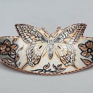 Op de afbeelding: Decoratieve metalen broche in de vorm van een vlinder. De broche heeft een gedetailleerd vlinderontwerp met bruin en zwarte vleugels, tegen een zilveren achtergrond. Bloemen- en bladeraccenten bevinden zich aan de zijkanten, met een koperkleurige rand.