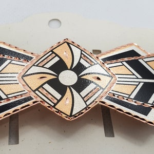 May include: A geometric patterned hair clip with a rose gold finish. The clip features a black, white, and peach design.