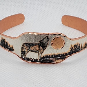 May include: A copper bracelet with a silver-toned wolf howling at the moon design. The moon is a copper color and the wolf is black and white.
