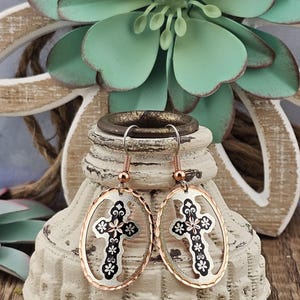 May include: A pair of silver and rose gold earrings with a cross design. The cross is surrounded by a floral pattern and is set in an oval shape.