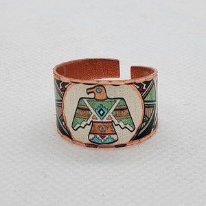 Pode incluir: Anel de cobre com design aberto, apresentando um design Thunderbird de inspiração nativa americana. O anel tem uma banda de cobre com uma imagem pintada de um pássaro em tons de laranja, turquesa, verde e preto. O anel tem aproximadamente 2.5 cm de largura.
