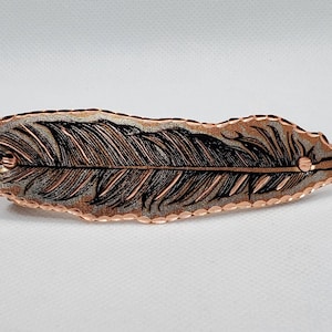 Puede incluir: Horquilla en forma de pluma de color cobre con detalles de plumas grabados. La horquilla tiene un borde texturizado y un centro plateado. El diseño de la pluma está detallado con líneas negras, creando un contraste llamativo. Un accesorio para el cabello único.