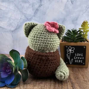 May include: A crocheted green cactus with a pink flower on top, sitting in a brown crocheted pot. The cactus is sitting on a wooden surface with a small succulent plant in a wooden planter next to it. The planter has a sign that says "Love Grows Here".
